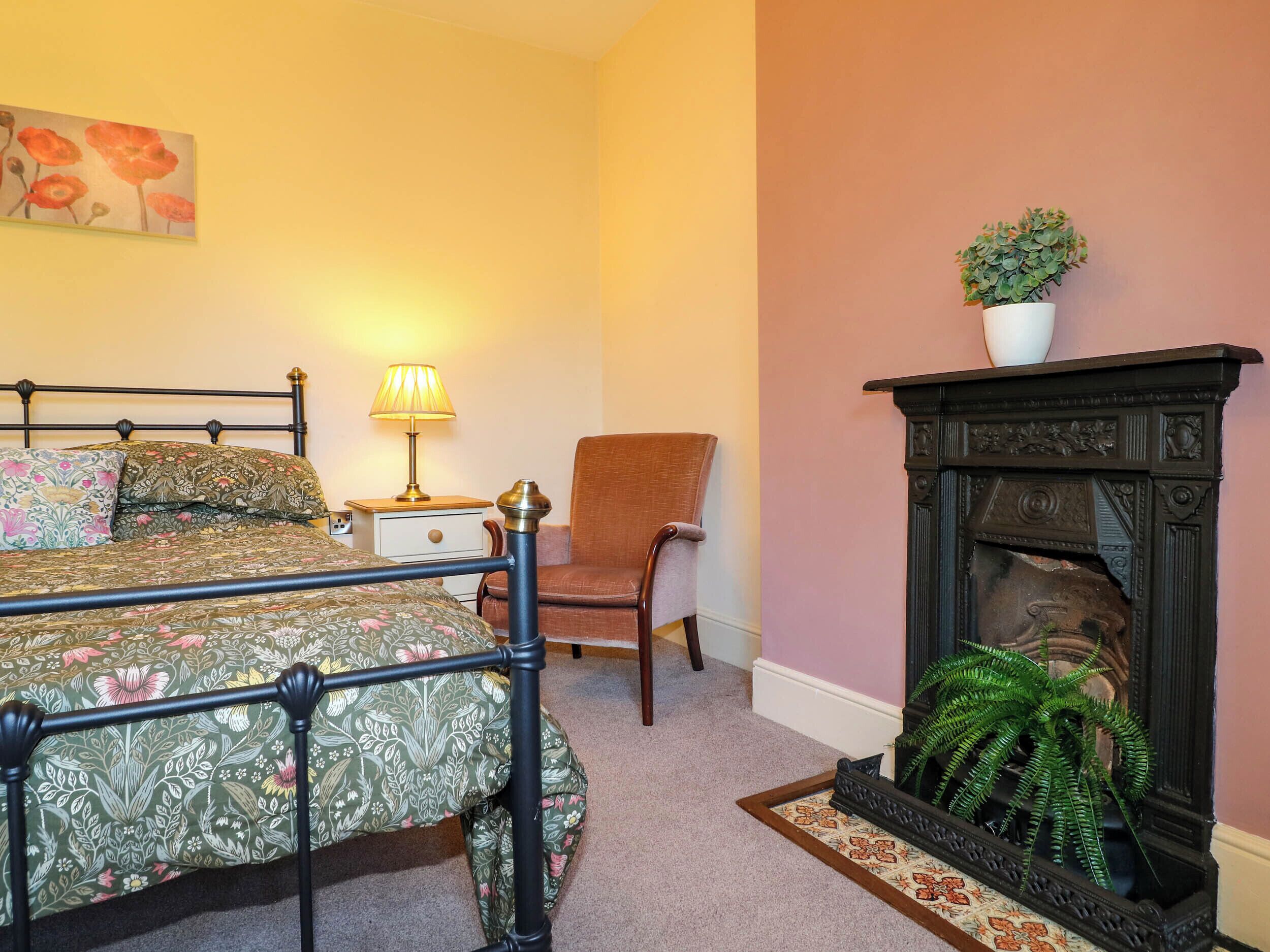 Ein Schlafzimmer mit Bett, Sessel, Nachttisch mit Lampe, Kamin und Blumentopf.
