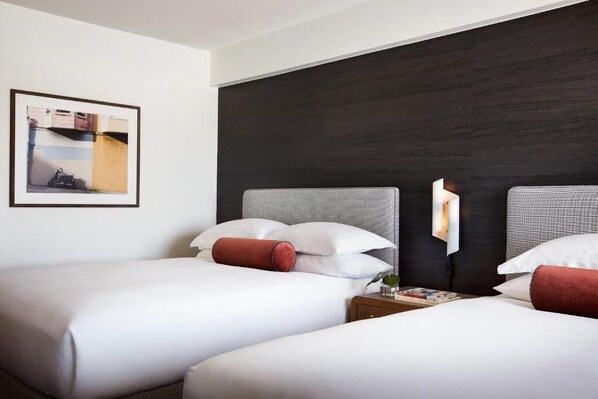 Habitación de hotel con dos camas, cabecero oscuro, un cuadro enmarcado en la pared y una mesita de noche con lámpara.
