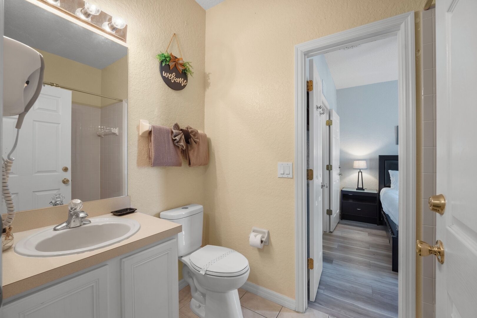 A bathroom with a sink, toilet, and mirror. There is a welcome sign on the wall and a towel rack with towels.