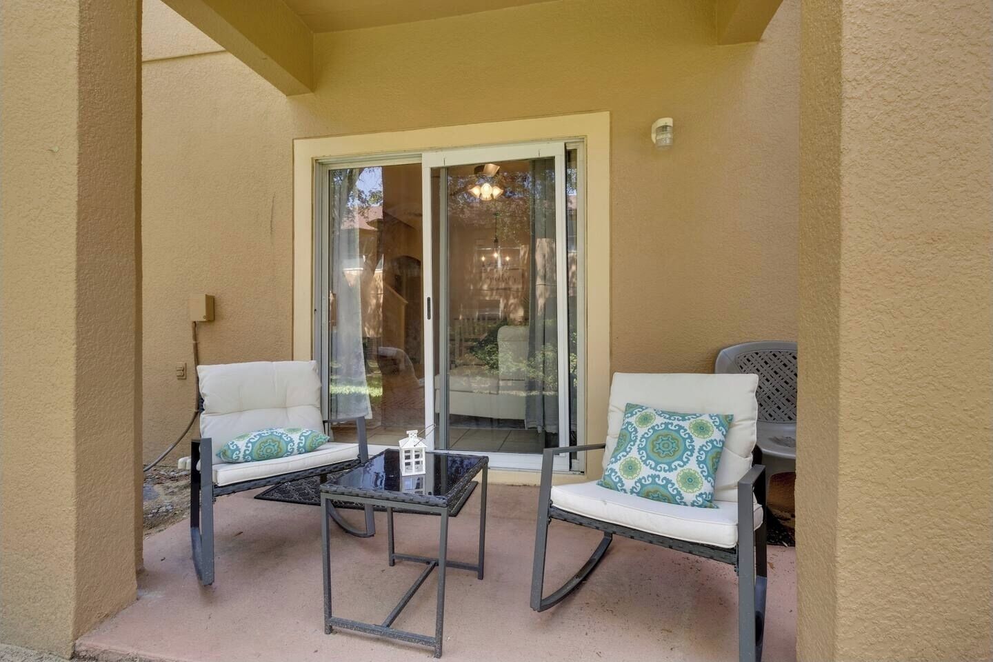 Un espacio de patio cubierto con dos sillas blancas acolchadas, una mesita y un ventanal de vidrio que da a un espacio al aire libre.