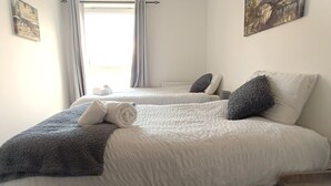 A hotel room with two beds, each with a white comforter and a dark throw blanket. There are pillows on the beds, and a window with curtains is visible.