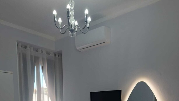 Ένα σύγχρονο υπνοδωμάτιο με ένα κρεβάτι, μια τηλεόραση τοποθετημένη στον τοίχο, ένα πολυέλαιο και μια μονάδα κλιματισμού.