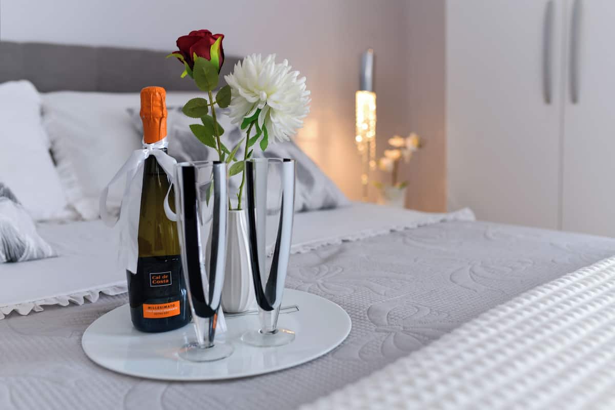 A bed with a white bedspread, a bottle of wine, and two vases with flowers on a bedside table.