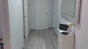 Ένας στενός διάδρομος με ανοιχτόχρωμο γκρι ξύλινο δάπεδο, λευκούς τοίχους και μια λευκή πόρτα στο τέλος. Στη δεξιά πλευρά υπάρχει ένα μικρό λευκό ντουλάπι με ένα βάζο με λουλούδια και έναν φορητό υπολογιστή.