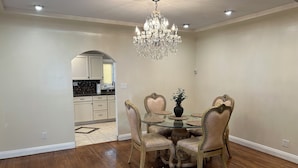 A dining area with a glass table surrounded by ornate chairs, a chandelier, and a view into the kitchen.
