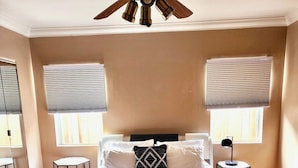 A bedroom with a large bed, two bedside tables, a ceiling fan, and two windows with blinds.