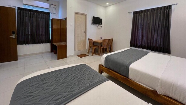 A hotel room with two beds, a ceiling fan, a TV, a desk with chairs, and a door. - Cholas Castle inn (Tiruchirappalli)