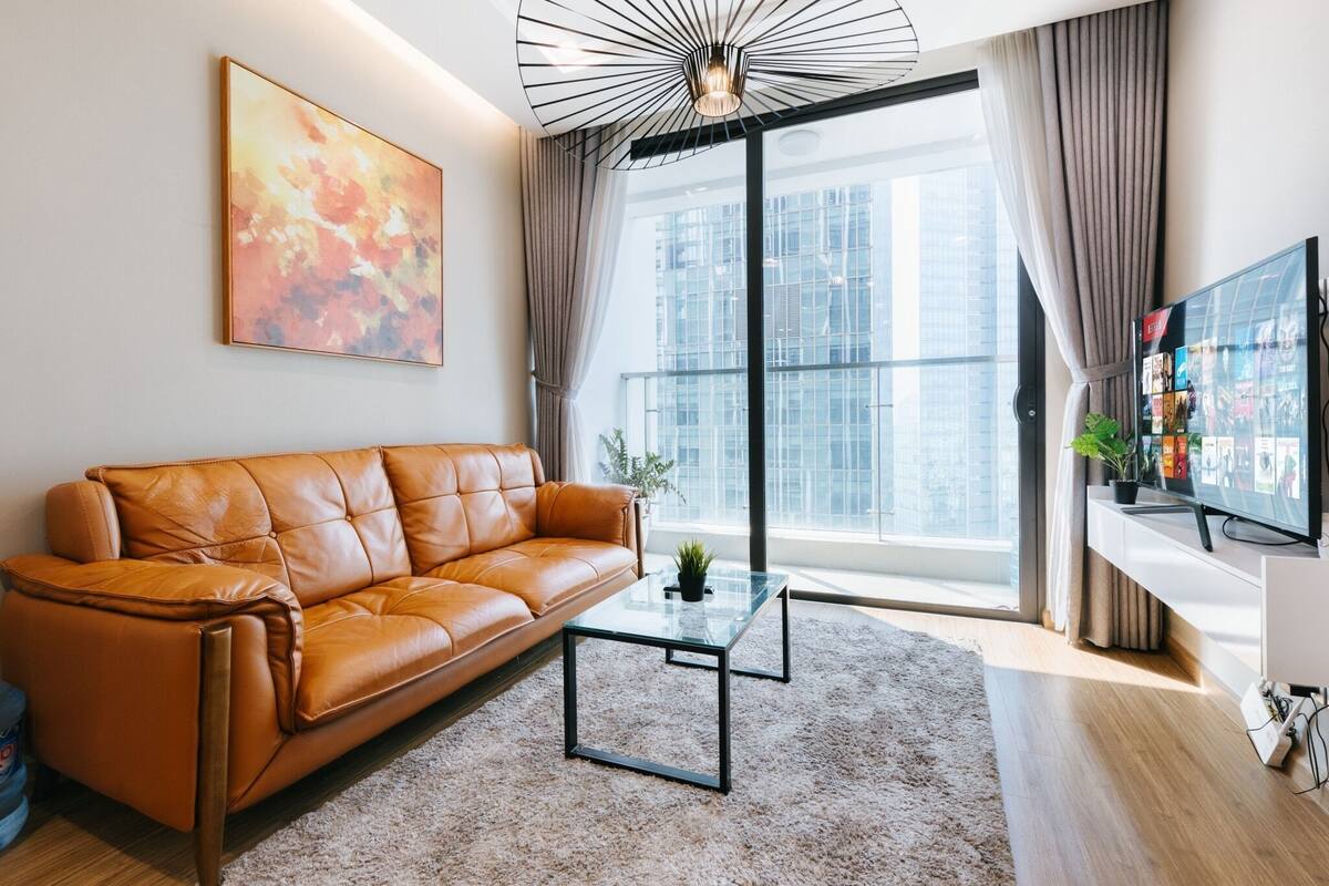 Ein modernes Wohnzimmer mit einer Ledersofa, einem Glastisch, einem großen Fenster mit Vorhängen und einem Flachbildfernseher auf einem weißen Schrank.