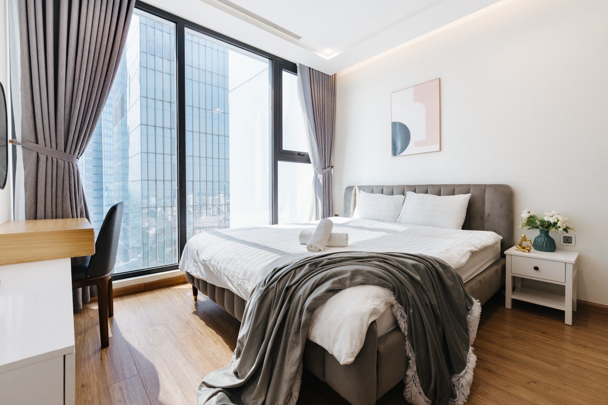 A modern hotel room with a large bed, a desk, and a view of a cityscape through large windows.