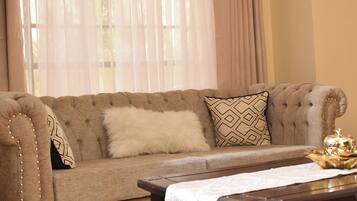 A tufted sofa with a fluffy white throw pillow and a patterned pillow, a wooden coffee table with a gold decorative item, and sheer curtains allowing natural light.