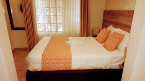 A hotel room with a bed, a window with curtains, and a pair of slippers on the floor. - The Marais Hotel and Suites- SIAYA (Siaya)