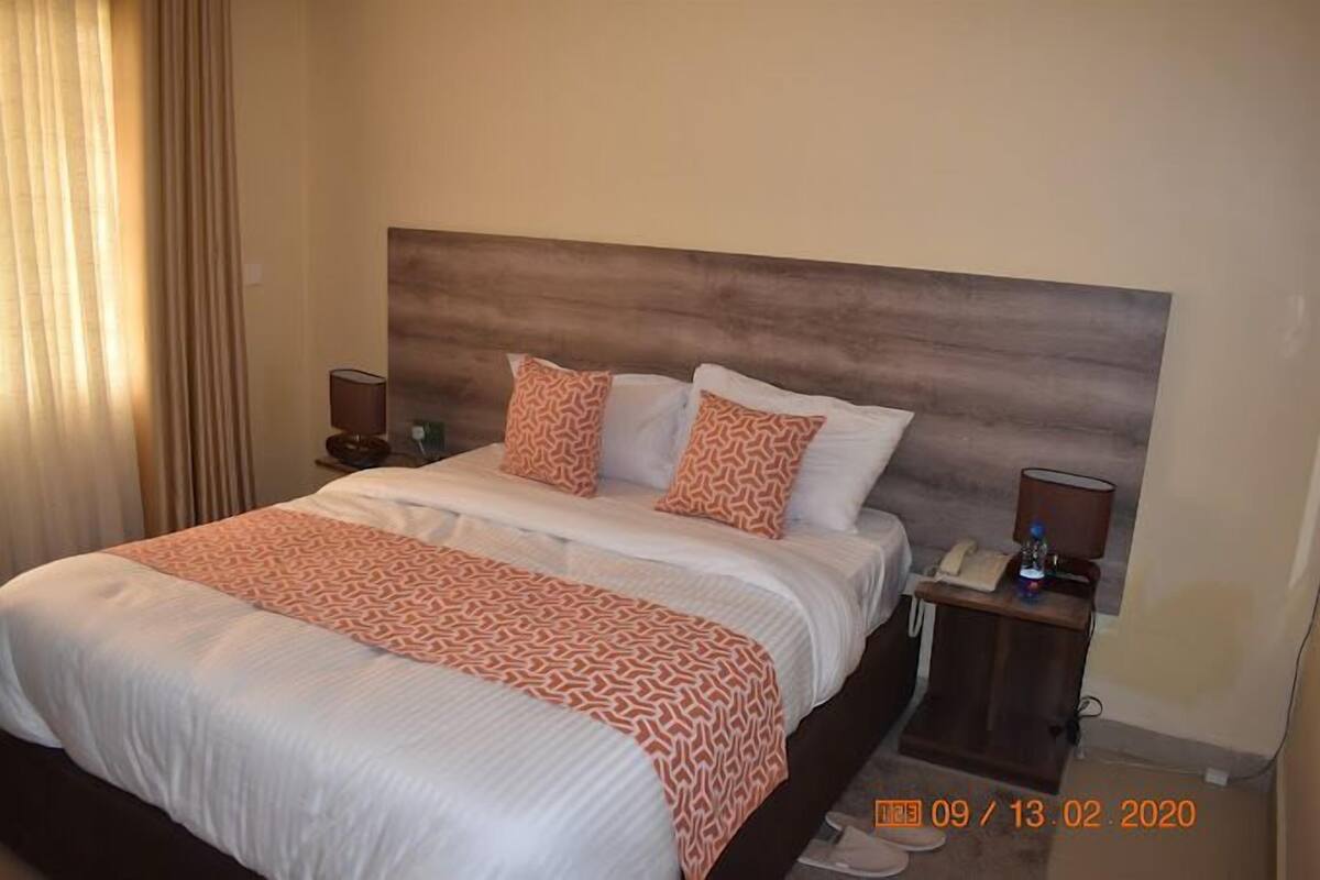 Una habitación de hotel con una cama grande, dos almohadas, una mesita de noche con una lámpara y un atril con un teléfono y una botella.