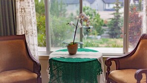 A room with a view of a garden through a window, a table with a lace tablecloth, and two wooden chairs. - Cozy tidy private | Sunset Garden (Cochrane)