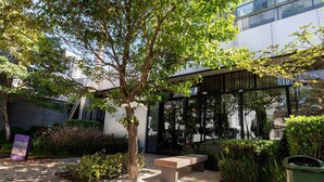 Un espacio de estar al aire libre con un banco, rodeado de follaje y un edificio con ventanas amplias.