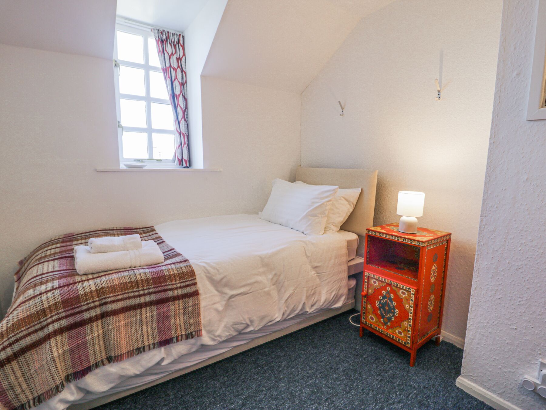 A bedroom with a bed, a window with curtains, a bedside table with a lamp, and a carpeted floor.