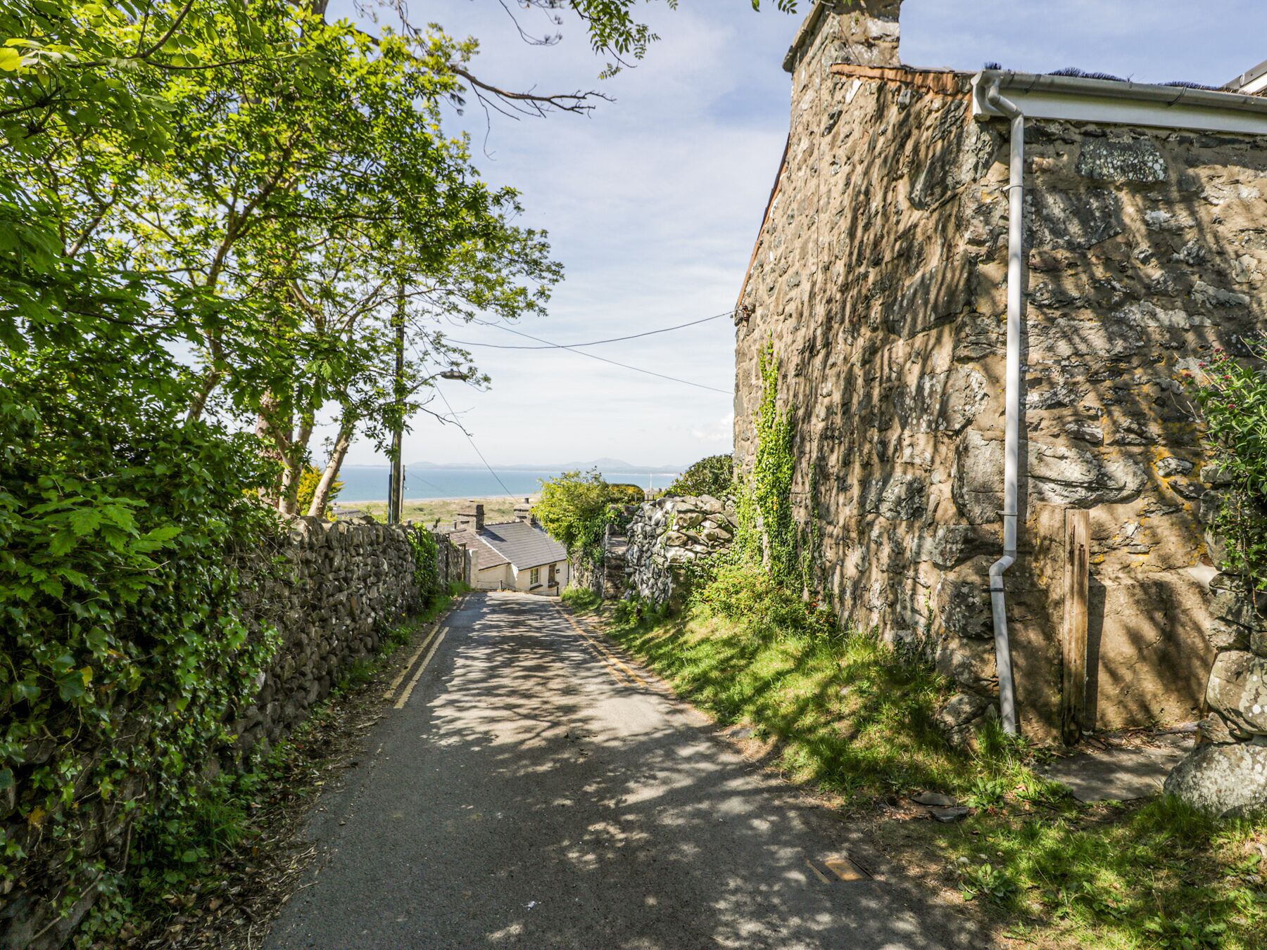 A narrow, paved road flanked by stone walls and lush greenery, leading to a stone building with a visible pipe.