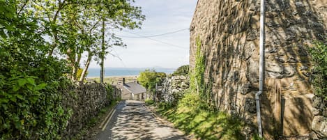 A narrow, paved road flanked by stone walls and lush greenery, leading to a stone building with a visible pipe.