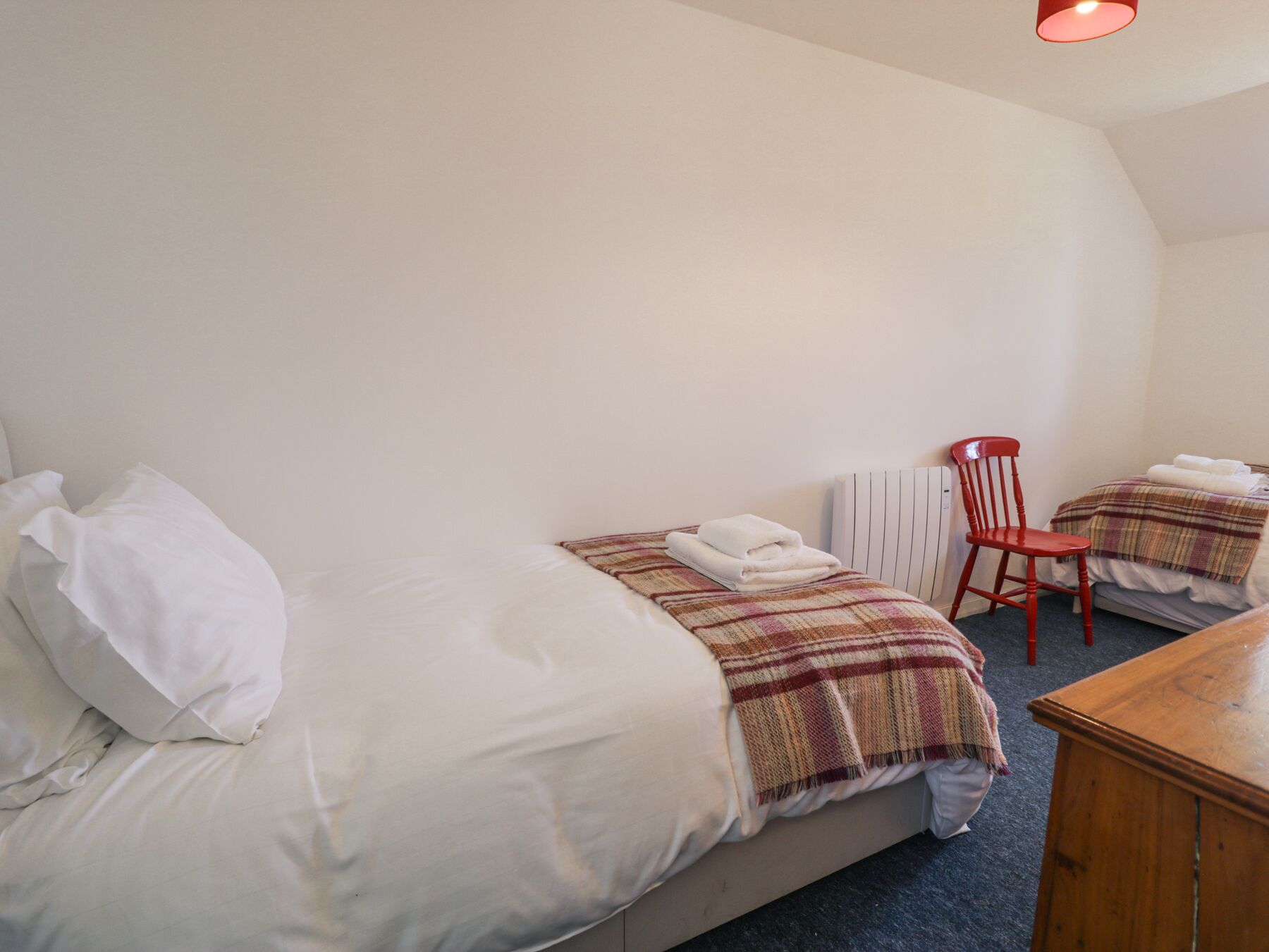 Una cama individual con ropa blanca, un plaid y una toalla doblada. Hay una silla roja y una mesa de madera.
