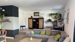 A modern living room with a grey sectional sofa, a wooden coffee table, and a dining area with a wooden table and chairs. There are shelves with plants and books, and a wooden cabinet.