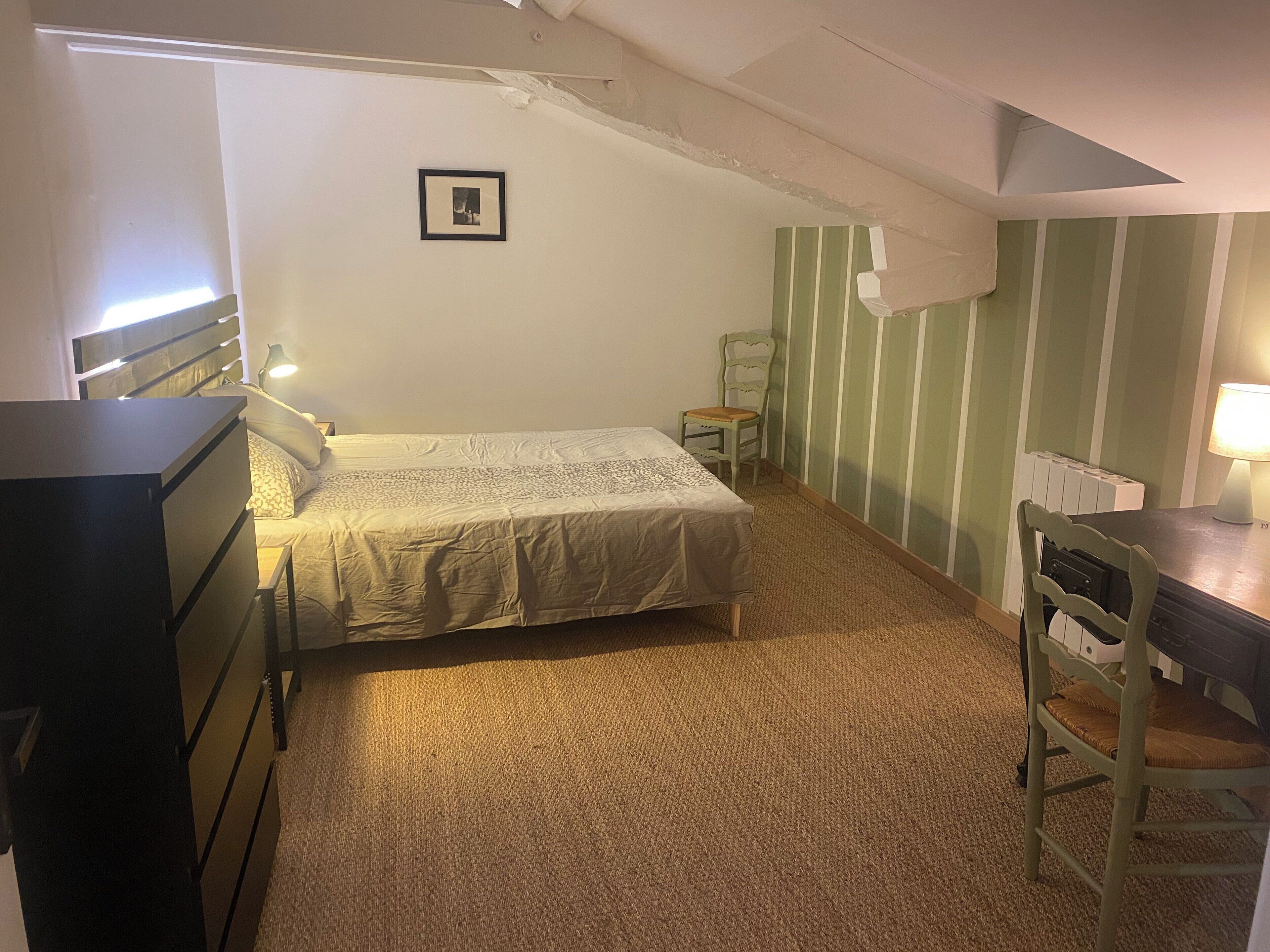Une petite pièce agréable et confortable, avec un lit, un bureau et une chaise. Il y a un radiateur au mur et un tableau accroché au-dessus du lit.