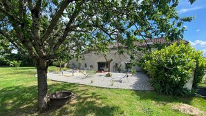 A rustic stone building with a tiled roof, surrounded by a gravel courtyard and lush greenery.