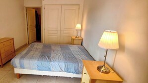 Un dormitorio con cama, mesita de noche con lámpara, cómoda y puerta que da a otra habitación.