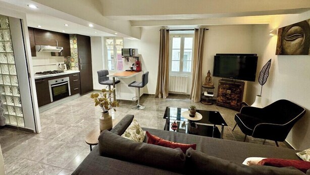 A modern living room with a sofa, a coffee table, and a TV. There is a kitchen area with a microwave and a stove.