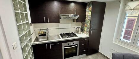 Una cocina compacta con armarios oscuros, una estufa a gas, un lavamanos y un ventanal con persianas.