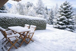 A snow-covered outdoor seating area with a table and chairs, a hedge, and snow-laden trees in the background.