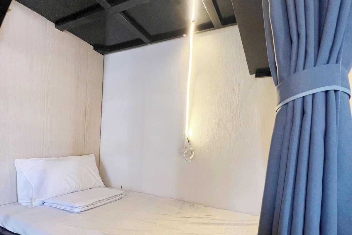 A single bed with white bedding, a white pillow, and a bedside table with a lamp.