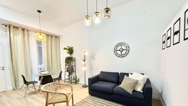 A modern living room with a dark sofa, wooden chairs, a striped rug, a round wall clock, and pendant lights.