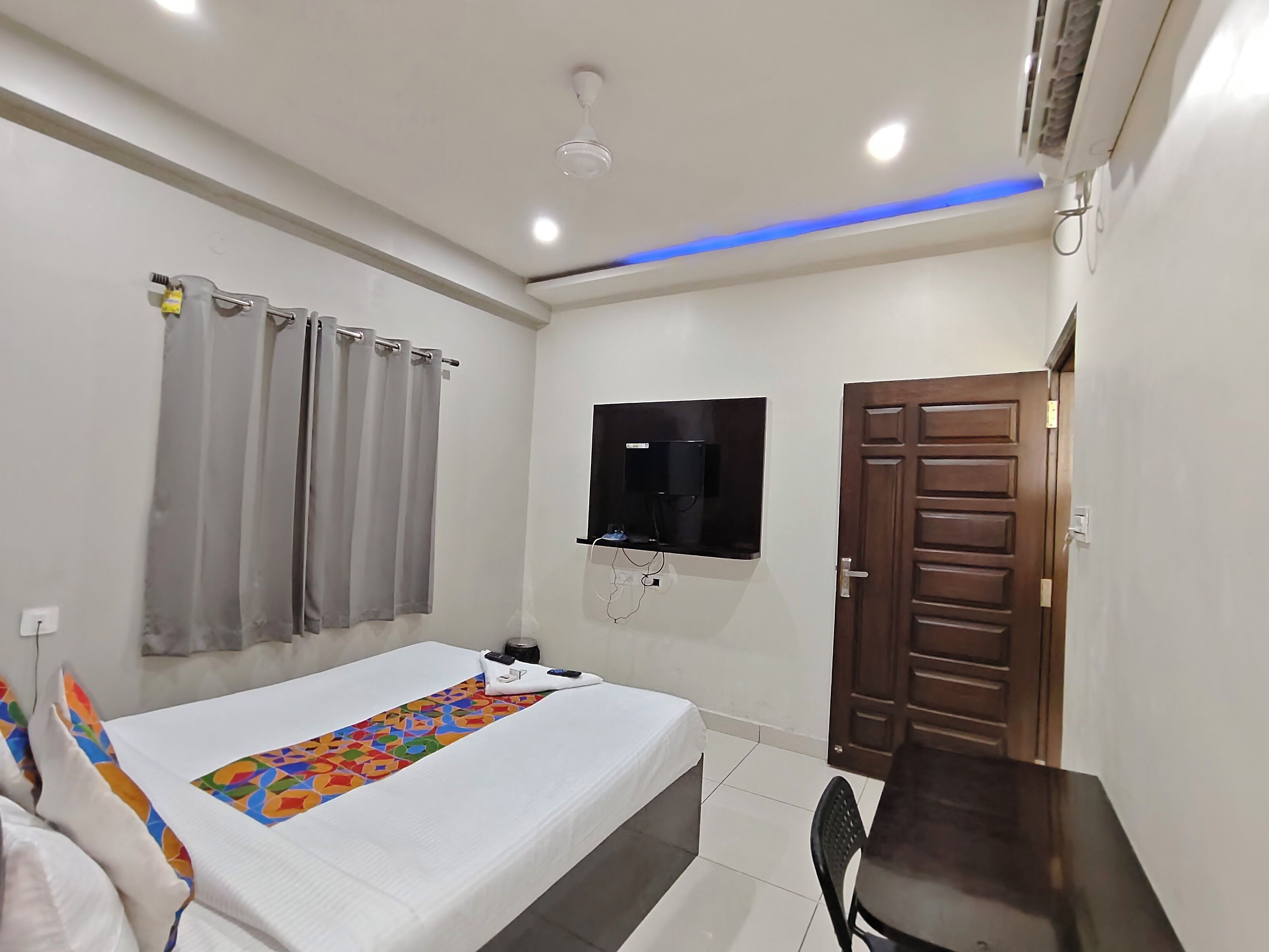 A bedroom with a bed, a TV mounted on the wall, a wooden door, and a ceiling fan.