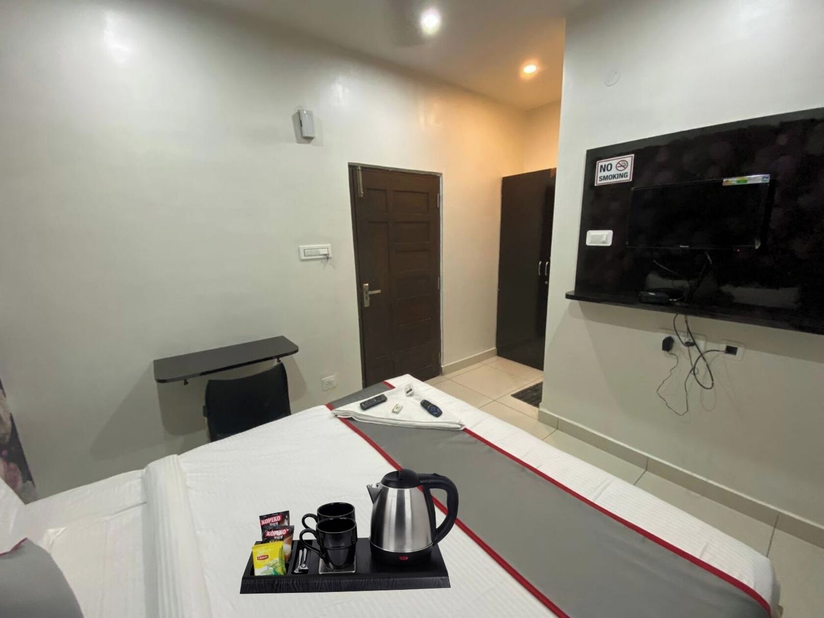 A hotel room with a bed, a small table with a kettle and cups, a TV mounted on the wall, and a door leading to another room.