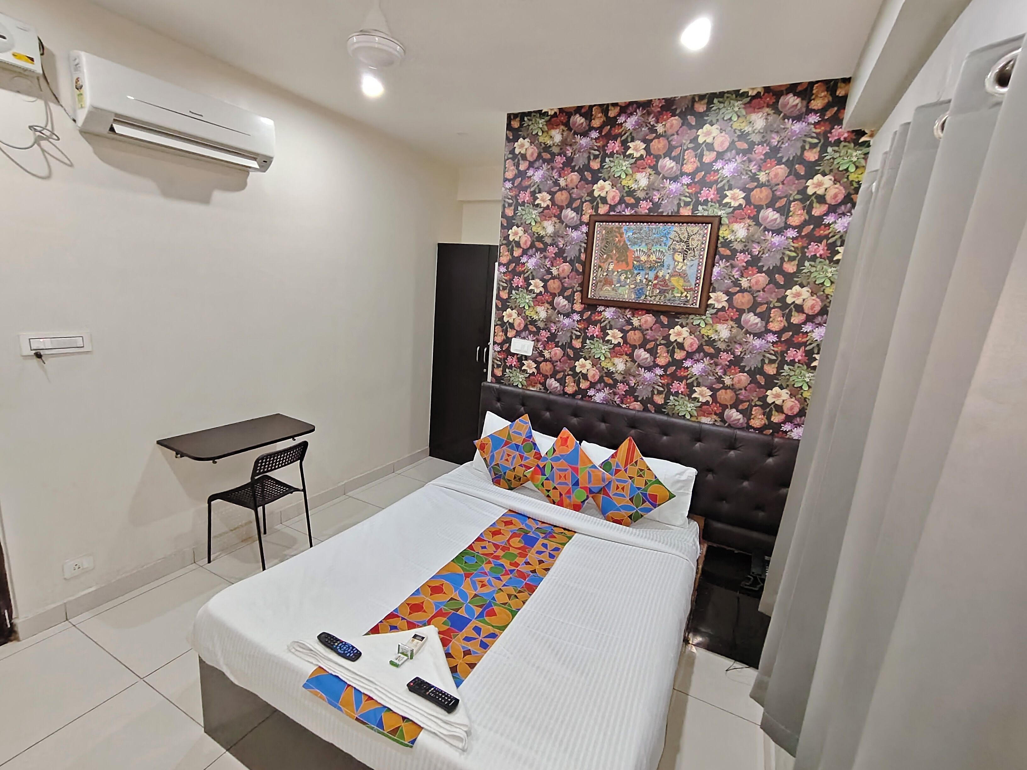 Een hotelkamer met een bed, een kleintje tafeltje, een stoel, een airconditioningapparaat, bloemenbehang en een ingelijst schilderij aan de muur.