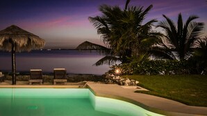Un área junto a la piscina con sillones de descanso y una sombrilla de paja, con vista a una masa de agua tranquila al atardecer.
