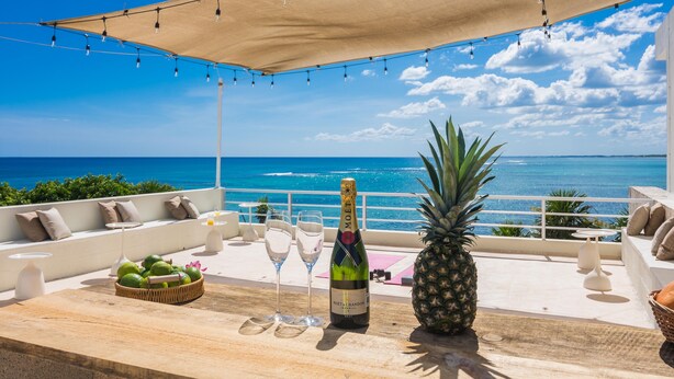 Una terraza con vista al océano, una botella de champán Moët, copas, una piña y una cesta de frutas.