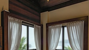 A bedroom with a large bed, two windows with curtains, a view of the sea, and a wooden ceiling. - Olhos verdes Guest house (Bombinhas)
