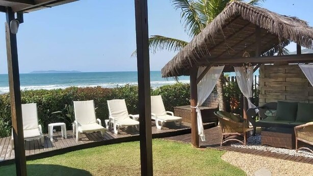 A beachside lounge with white lounge chairs, a thatched-roof gazebo, and a view of the ocean. - Olhos verdes Guest house (Bombinhas)