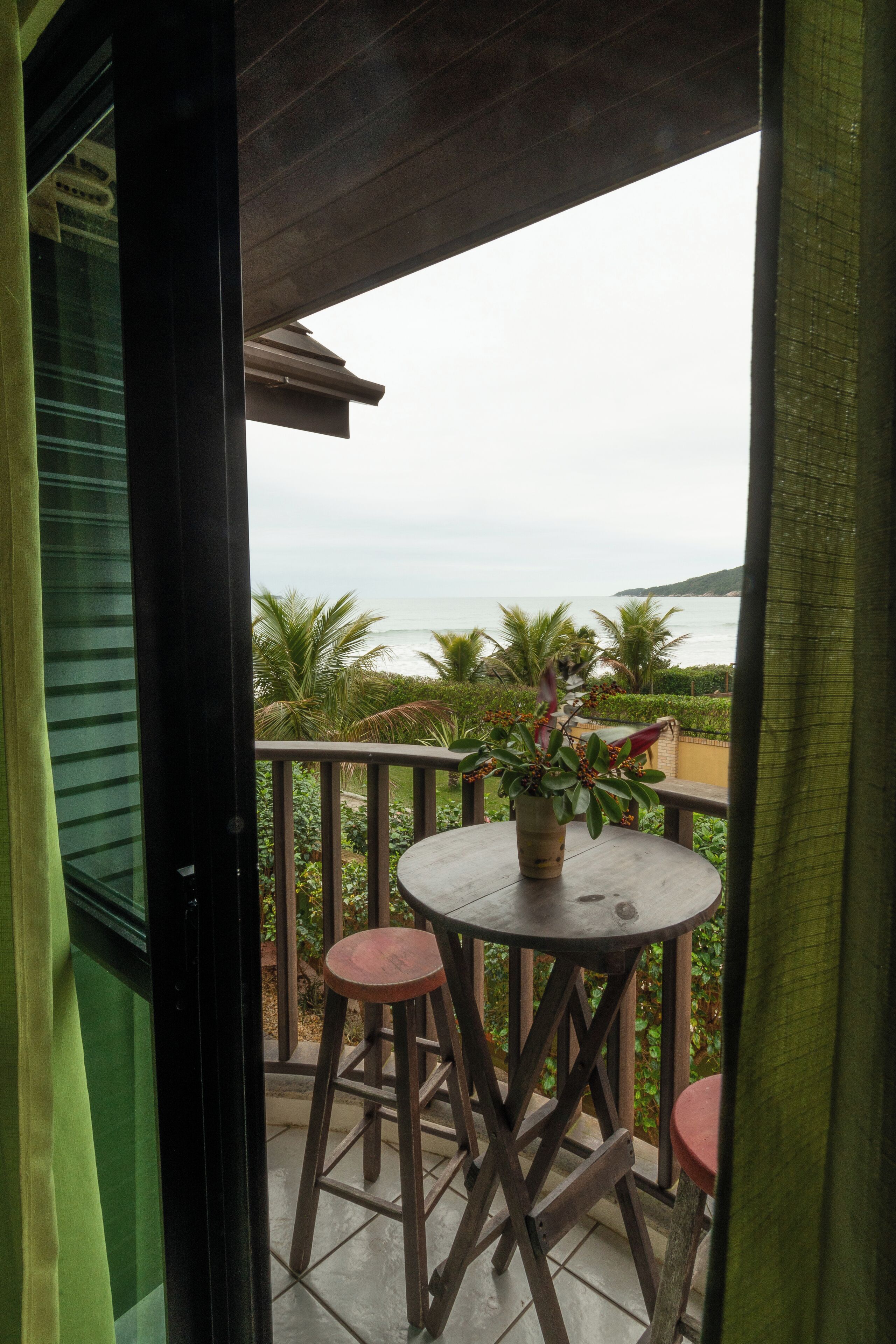 Ein Balkon mit rundem Tisch und zwei Hocker, der auf eine Küstenlandschaft mit Palmen und dem Meer blickt.