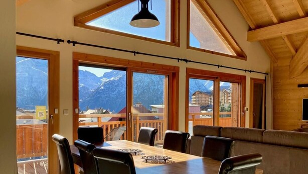A dining area with a wooden table and chairs, large windows with mountain views, and a balcony with a wooden railing. - 250m from ski slopes, 6th floor, balcony, tv, 99m², Les 2 Alpes (Les Deux Alpes)