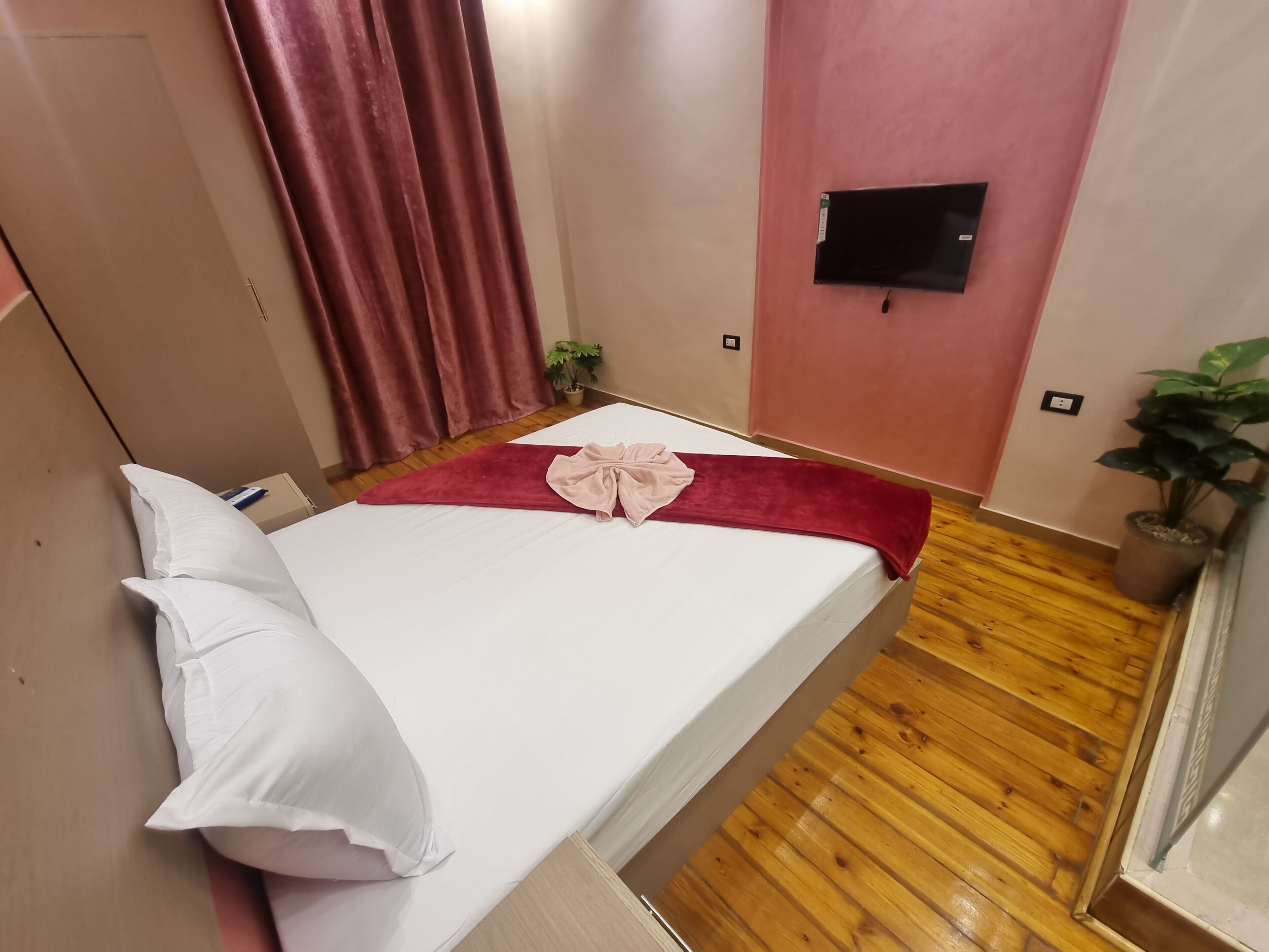 A single bed with white linens, a red blanket, and a pink towel. There is a potted plant on the floor and a TV mounted on the wall.
