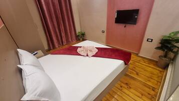 A single bed with white linens, a red blanket, and a pink towel. There is a potted plant on the floor and a TV mounted on the wall.
