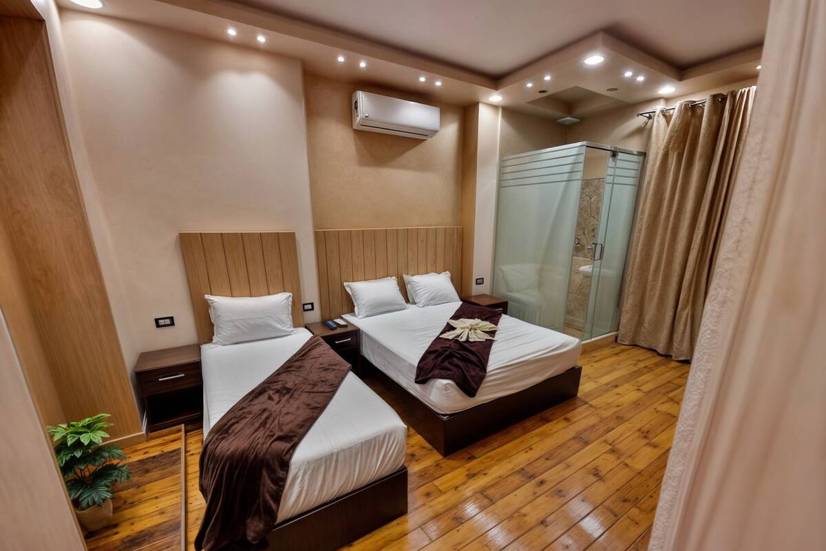 A hotel room with two beds, wooden flooring, an air conditioning unit, a glass shower enclosure, and a potted plant.