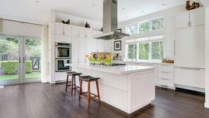 Een moderne keuken met witte kastjes, een centraal eiland met krukken, dubbele ovens en een groot raam dat voor natuurlijk licht zorgt.