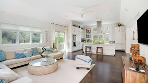 Een ruime woonkamer met een bank, een ronde salontafel en een keukenhoek met witte kasten en een groot raam.