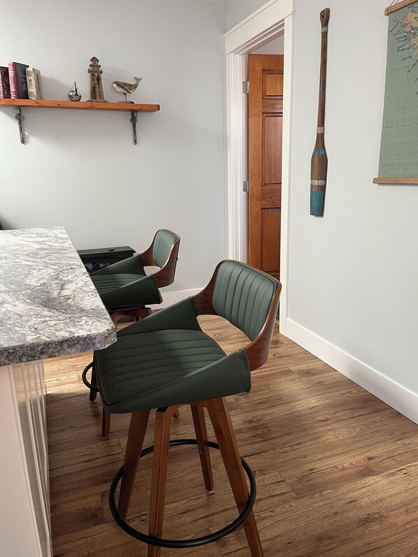 A dining area with two green upholstered bar stools, a wooden table, and a shelf with books and decorative items. - Steps to the Lake and Hiking Trails. Central Home Base for Island Adventures! (Nanaimo)