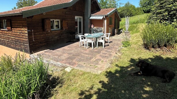A wooden house with a tiled roof, a patio with a table and chairs, and a dog lying on the grass. - Nature chalet on 8000m2 of totally quiet land in Coinchimont (Ban-de-Laveline)