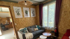 A cozy living room with a sofa, a small round table, and a red couch. There are framed artworks on the wall and a window with curtains. - Nature chalet on 8000m2 of totally quiet land in Coinchimont (Ban-de-Laveline)