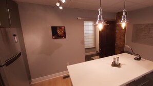 Een moderne keuken met een wit aanrecht, hangende hanglampen, een koelkast en een schilderij aan de muur.
