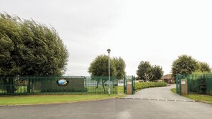 A gated entrance with a sign, a paved road, and trees. - Sweetacres (Great Yarmouth)